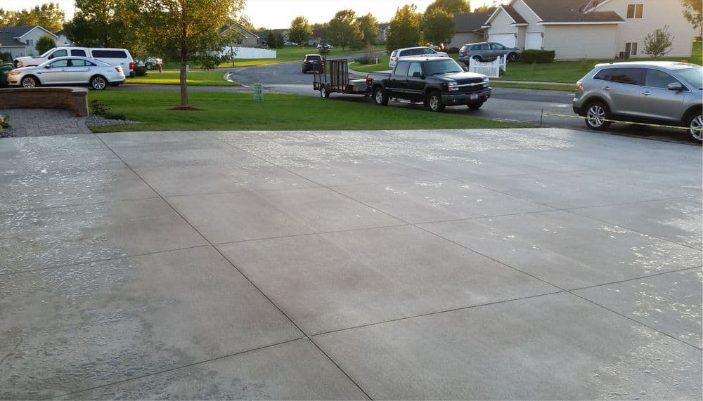 Newly poured concrete driveway with two parked cars and a trailer in a suburban neighborhood.