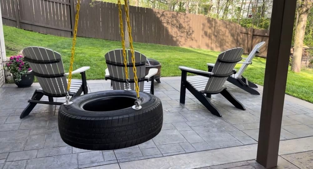 Tire swing hanging near patio chairs in a landscaped backyard setting.