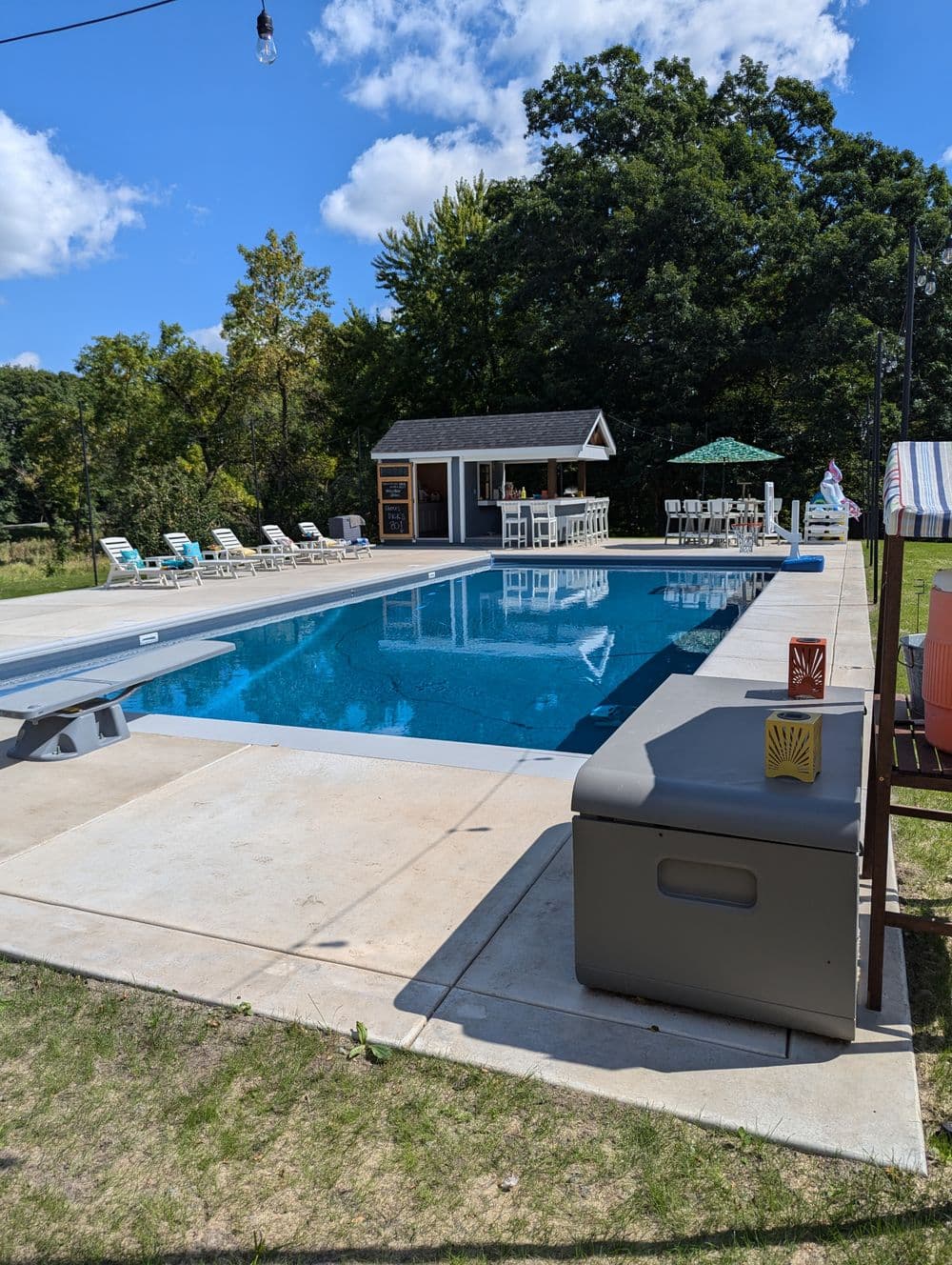 Swimming pool with lounge chairs and bar area surrounded by trees under a blue sky.