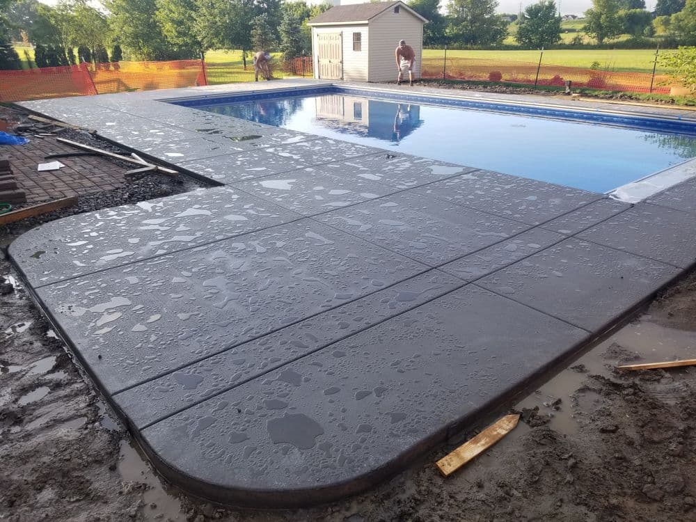 Newly completed pool deck surrounding an in-ground swimming pool in a landscaped yard.