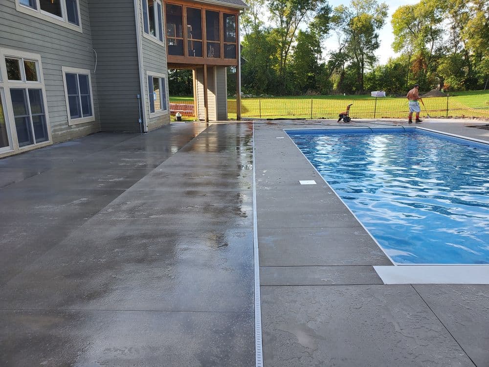 Concrete poolside area with a clear blue swimming pool and a house in the background.