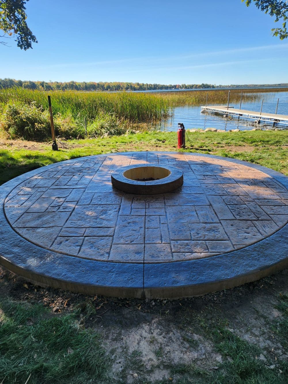 Circular stone fire pit surrounded by a grassy area near a serene lake.