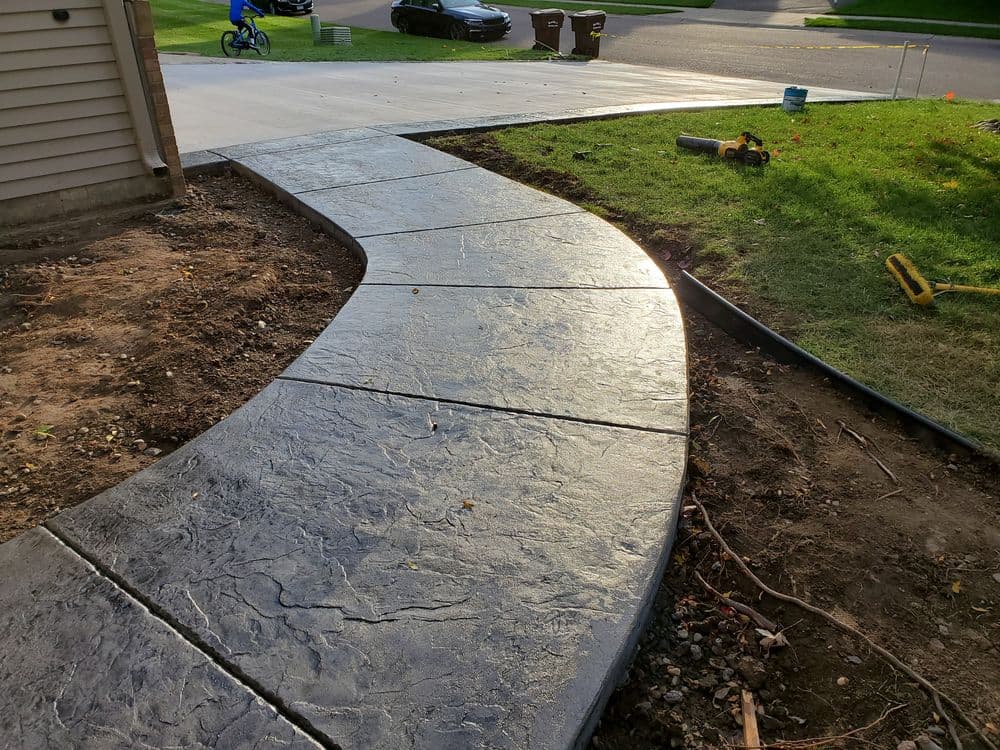 Curved stamped concrete pathway leading to a driveway with grass and landscaping details.
