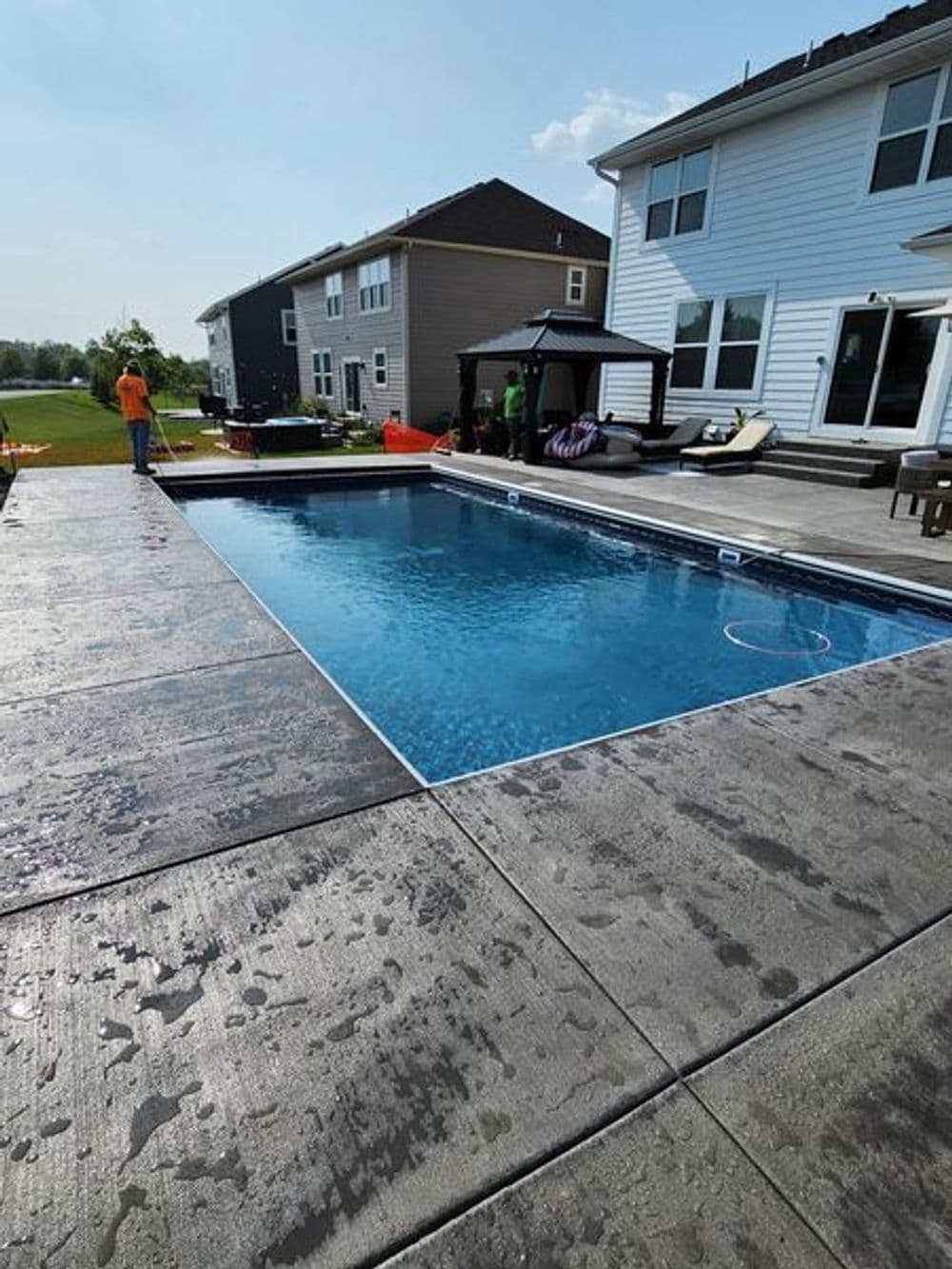 Backyard swimming pool with patio area, lounge chairs, and sunny sky in residential setting.