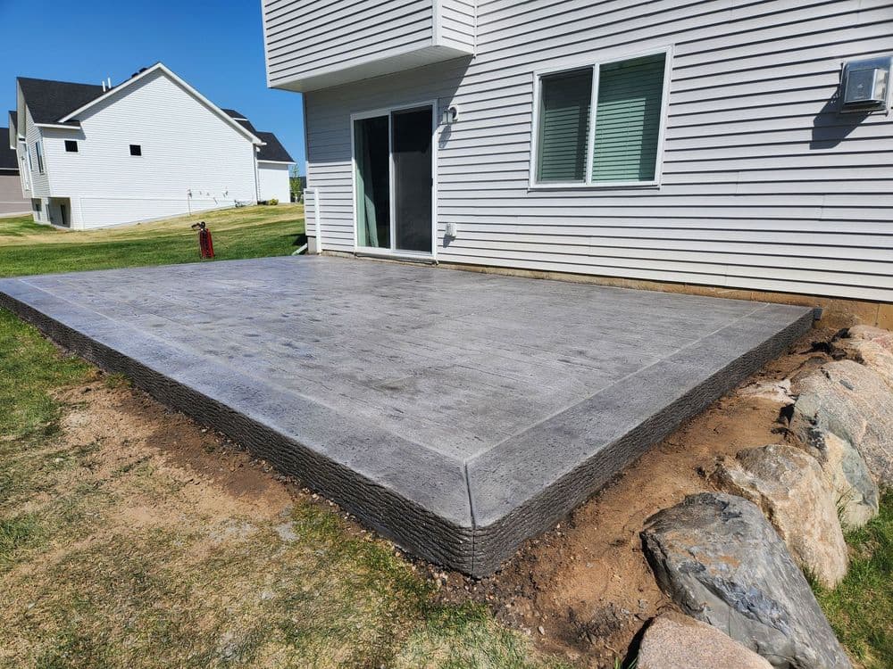 Newly poured concrete patio beside a house, featuring trimmed grass and rock edging.
