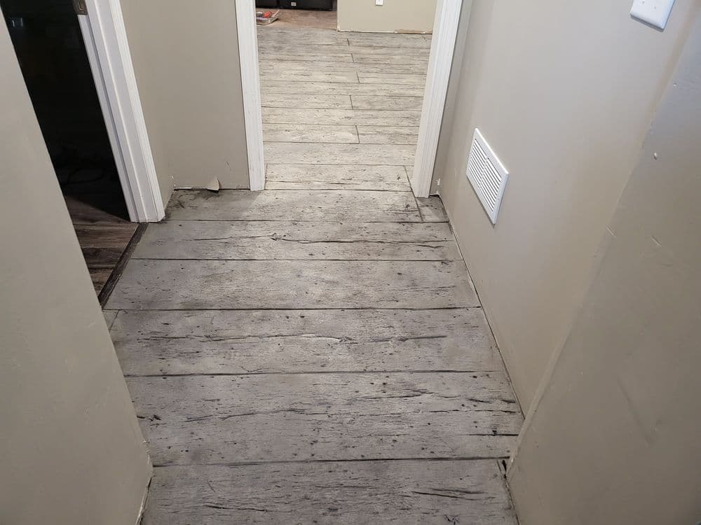 A hallway with gray wooden flooring and light walls, leading to another room.