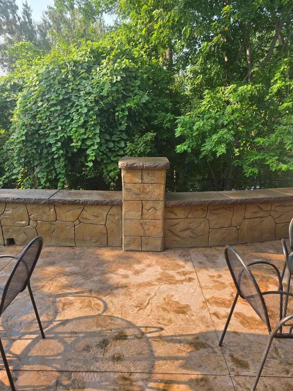 Stone wall with greenery in the background and patio chairs on a sunny day.