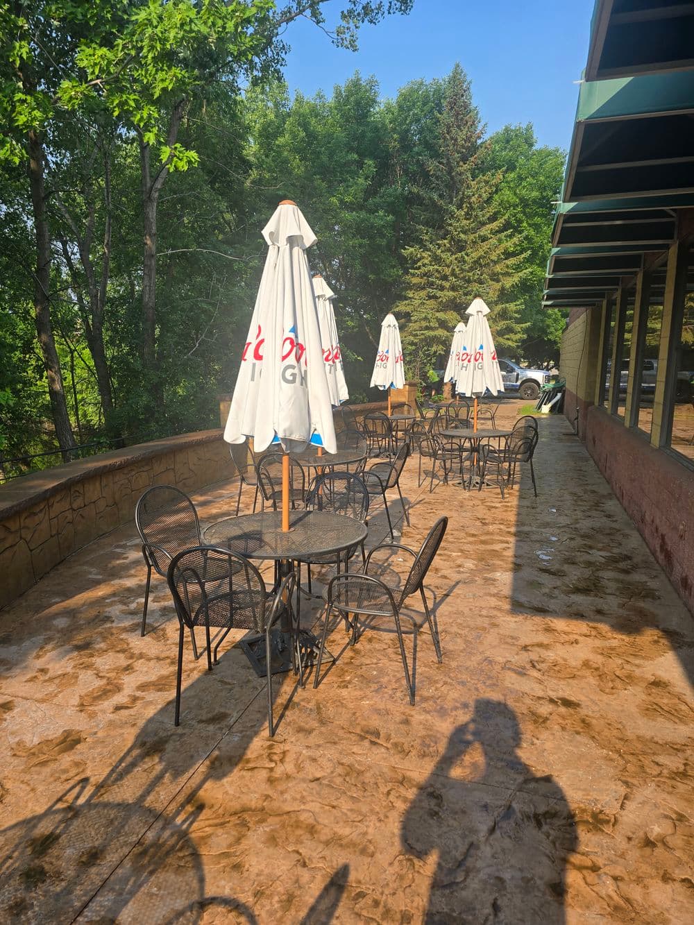 Outdoor café patio with tables and umbrellas surrounded by trees on a sunny day.